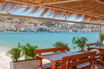 Beautiful summer landscape of sea resort – café on the beach: wooden tables and benches, flowers and cityscape on background
