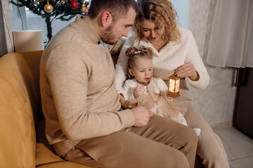 Young parents with their one-year-old daughter at home near the Christmas tree celebrate the Christmas holiday