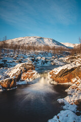 Winter fairy tale in the form of Storforsen waterfall in Silsand, Senja, Norway. Sunset illuminates the wild waterfall. In the background, the snowy hills of the Norwegian landscape