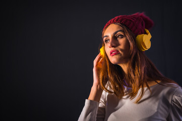 Portrait of a young brunette Caucasian woman with yellow headphones, a white T-shirt and a woolen hat on a black background, concentrated listening to music