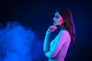 Portrait of a young brunette Caucasian woman in a white t-shirt dancing a disco with a black background. Neon lights with a background smoke