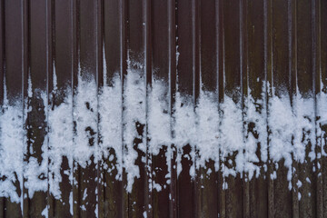 Fragment of a brown metal fence. A white, uneven strip of snow adhered to the surface of the profiled sheet. Background. Texture.