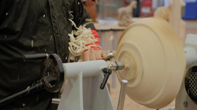 Man carpenter using chisel for shaping piece of wood on turning lathe machine with many shavings, making wooden bowl at workshop - close up. Carpentry, woodworking and manufacturing concept