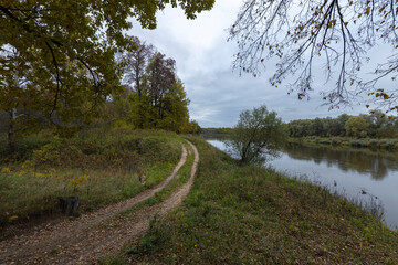 Obraz premium Dirt road along the river bank. Cloudy autumn weather. Rural landscape with road, river and autumn forest.