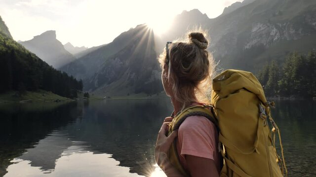 Young woman hiking by the lake, stunning mountain view. Female hiker walks at the edge of an Alpine lake. Sun is setting beyond the Alpine peaks