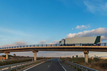 Truck with refrigerated semi-trailer driving on a bridge that crosses a highway.