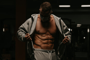 Fototapeta na wymiar A bodybuilder in a face mask is opening his hoodie to demonstrate his abs in gym