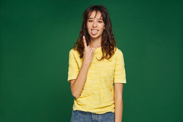 Modern cool young girl show rock-n-roll gesture, stick tongue, female do devil horns hand sign on green background