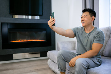 a young cheerful Asian sits on the couch at home and communicates during a video call or takes a selfie on a mobile phone