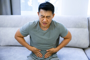Asian man suffering from abdominal pain, sitting on the couch in his living room. Symptoms of poisoning, diarrhea and coronavirus disease.