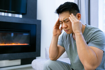 A tired Asian man is sitting on the sofa, he has a headache. Unhealthy guy has a cold or flu, is upset about an illness, unhappy man feels depressed