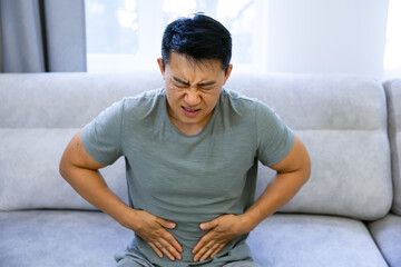 asian frustrated unhealthy guy, sitting at home on the sofa, holding his stomach with his hands,...