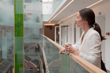 Fototapeta na wymiar young business woman in a white jacket is standing in a public space with a takeaway coffee in her hand. Confident brunette freelancer, portrait on the background of a business center.