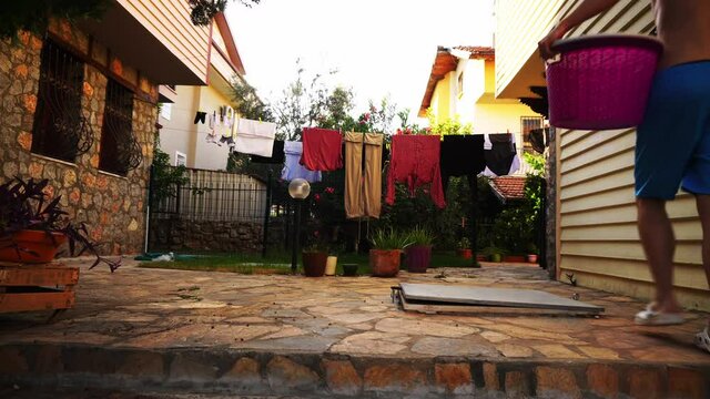 Man in blue shorts is taking off clothes from the rope which were drying after laundry outdoors. Young caucasian person is going with backet and picking pants and shirts from clothespin on the street.