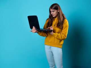 Online training, a young woman uses a laptop. A student with a laptop in her hands, a young brunette in a yellow hoodie on a blue background