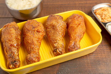 Deep Fried Chicken Drumsticks coated with hot and garlic sauce in Korean style served on bright yellow plate with blurred background of side dishes on wooden table