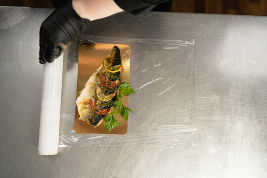 close-up of female hands in the kitchen in a restaurant pack the baked mackerel in plastic wrap.