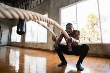 African sport man doing battle ropes functional training at gym. Determined trainer making waves with ropes while exercising strength. Athlete working out with battle rope at industrial gym.