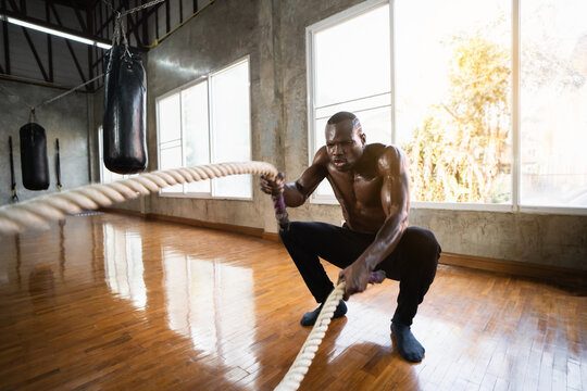 African sport man doing battle ropes functional training at gym. Determined trainer making waves with ropes while exercising strength. Athlete working out with battle rope at industrial gym.