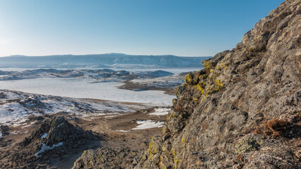 The frozen lake is surrounded by snow-covered hills. A mountain range against a blue sky. Granite rock with yellow lichens in the foreground. Baikal. Olkhon Island