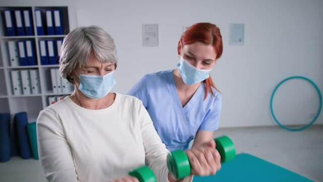 Home Health Care During Covid 19, Old Woman With Nurse In Medical Masks, Physiotherapist Helps Active Grandmother With Dumbbells