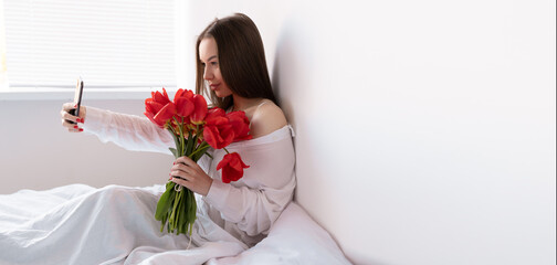 Portrait of a sexy girl with a bouquet of red tulips in the morning in the bedroom for Valentine's Day makes a selfie on her smartphone. Woman with flowers on the bed Women's day.