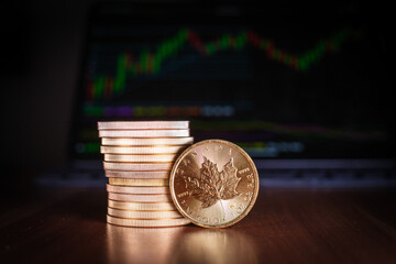 a Maple leaf gold coin stands in front of a stack of gold coins, in the background is a trading chart
