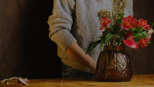 Unrecognizable woman makes flower arrangement using fresh and dried flowers. Florist cuts stems of plants, puts them in vase. Red and golden floral composition in vase.