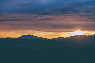 Atmospheric landscape with silhouettes of mountains with trees on background of dawn sky with sun circle and orange sun rays. Colorful nature scenery with sunset or sunrise of illuminating color.