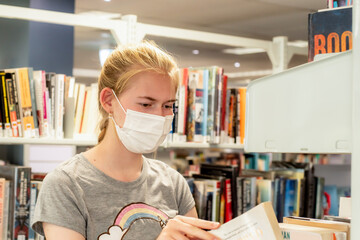 Teenage girl wearing face mask with the book in her hands at the library during the COVID-19...