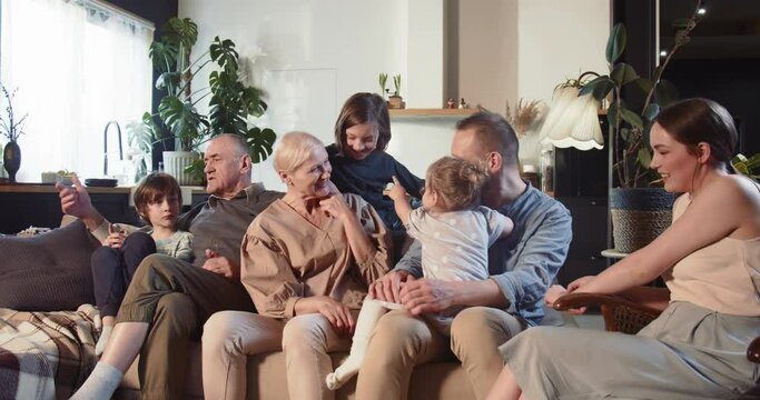 MULTI GENERATIONAL FAMILY Cheerful senior grandparents, young mom and dad, teens and baby girl talk on couch at home.