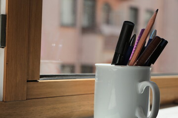 Pens in the white cup in front of the window. Felt-tip pen,ball-point pen, lead pencil, waterproof marker. - obrazy, fototapety, plakaty