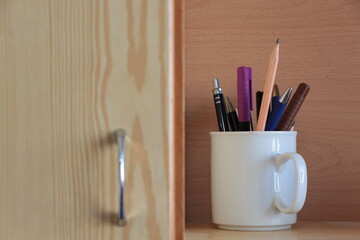 Obraz premium Pens in the white cup. Felt-tip pen,ball-point pen, lead pencil, waterproof marker. Wood in background.