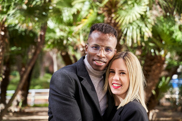 Fototapeta premium Black man and blond woman looking at camera while standing against palms during romantic date on sunny day in exotic park