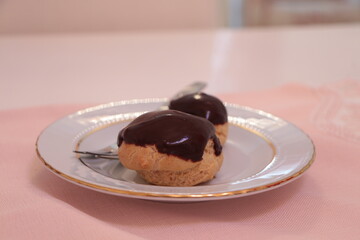 Homemade profiteroles covered with chocolate sauce on the pink tablecloth. Profiterole choux with chocolate ganache on the plate. Delicious eclair dessert with cream and chocolate glaze. 
