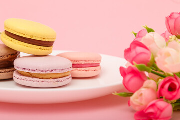 Fototapeta na wymiar Plate of macaroons on pink background with flowers. Sweet pastry, baked products, sweets, dessert. Unhealthy diet, sugar dependence.
