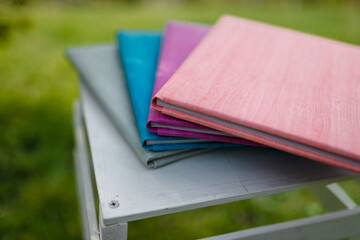 Spines of books, albums, notebooks or photobooks with grey, turquoise, purple and pink leather covers lying on white wooden box among green grass. Soft focused shot.