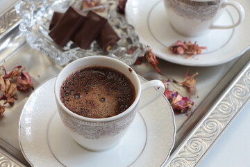 A white cup of Turkish coffee on silver tray.Traditional Turkish Coffee.Coffee foam.Small porcelain coffee cup with silver detail.Dried pink flowers and chocolates on a glass plate. Selective focus