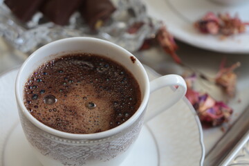 A white cup of Turkish coffee on silver tray.Traditional Turkish Coffee.Coffee foam.Small porcelain coffee cup with silver detail.Dried pink flowers and chocolates on a glass plate. Selective focus