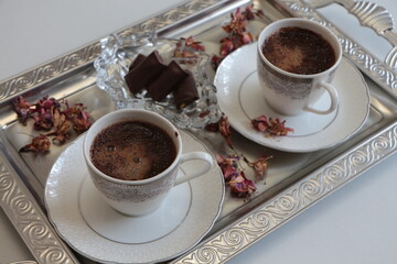 Two white cup of Turkish coffee on silver tray.Traditional Turkish Coffee.Coffee foam.Small porcelain coffee cup with silver detail.Dried pink flowers and chocolates on a glass plate. Selective focus