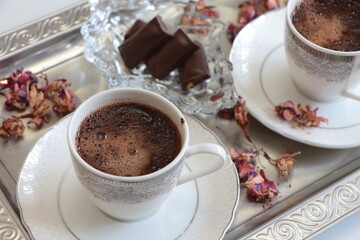 A white cup of Turkish coffee on silver tray.Traditional Turkish Coffee.Coffee foam.Small porcelain coffee cup with silver detail.Dried pink flowers and chocolates on a glass plate. Selective focus