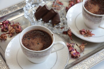 A white cup of Turkish coffee on silver tray.Traditional Turkish Coffee.Coffee foam.Small porcelain coffee cup with silver detail.Dried pink flowers and chocolates on a glass plate. Selective focus