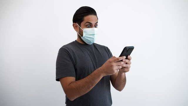 Portrait of caucasian man raising eyebrows because of surprise with smartphone and medical mask during corona virus outbreak. Surprised reaction person with phone device on white background