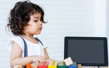 Caucasian adorable cute curly hair little girl wearing white shirt, staying alone, playing toys and tablet in living room at home with fun. Education and Kid Concept.