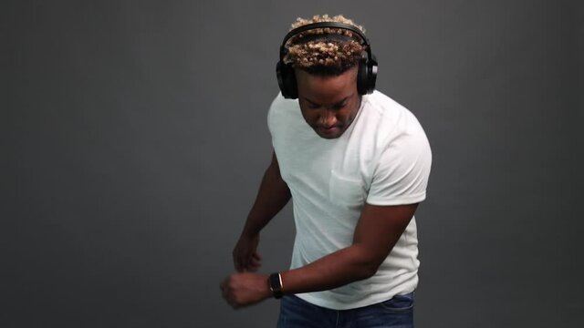 African guy in a white T-shirt listens to music with black headphones and dances on a gray studio background. Charismatic man with an African hairstyle lights up in the dance. 4k resolution