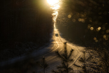 Obraz premium Sun rays through the trees and ice covered road with single car on it during the winter.