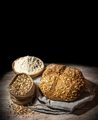 Loaf of cereal bread on wooden table and dark background. Wooden spoon and bowl decoration with flour and multigrain
