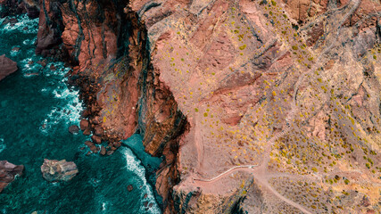 Atlantic ocean coastline with red rocky cliff, hiking path, turquoise waves and sea foam. Madeira island, Portugal. Aerial photography from drone. Breathtaking landscape.