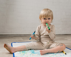 A little cheerful girl in pajamas is sitting on the floor and gnawing on water marker. The face and clothes are stained with paint
