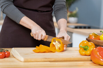Hands of young female chopping fresh yellow capsicum on wooden board while going to prepare...